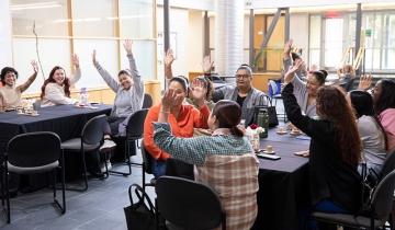 11 students sitting around tables enthusiastically raising their hands